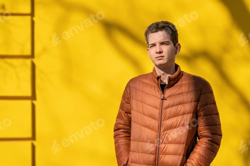 Preview: Portrait of a young man in a brown jacket stands on the street against a yellow wall