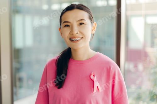 Preview: Young asian woman smiling and showing pink ribbon for breast cancer awareness