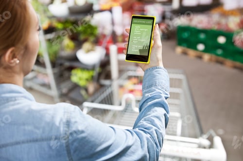 Preview: Woman holding a smart phone with a shopping list in front of a super market