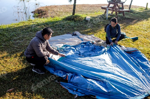 Preview: Two Men Kneeling on the Grass Working Together to Unfold a Blue Tent by the Side of a Lake