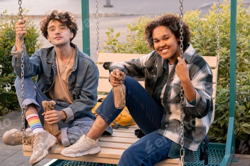 Preview: Happy restful teens having drinks while sitting on swings