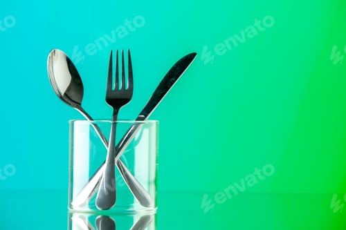 Preview: front view metal cutlery fork knife and spoon inside little glass on blue-green background color