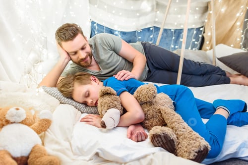 Preview: Smiling father looking at cute little son sleeping with teddy bear in blanket fort