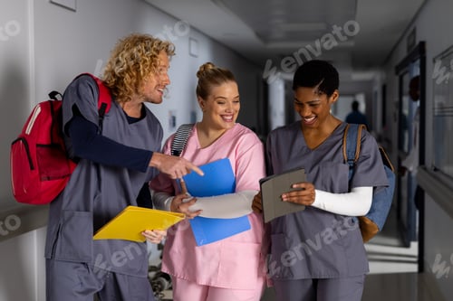 Preview: Happy diverse doctors wearing scrubs discussing work and using tablet in corridor at hospital