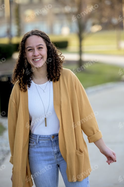 Preview: Woman with curly hair wearing a yellow jacket in a park under sunlight with a blurry background