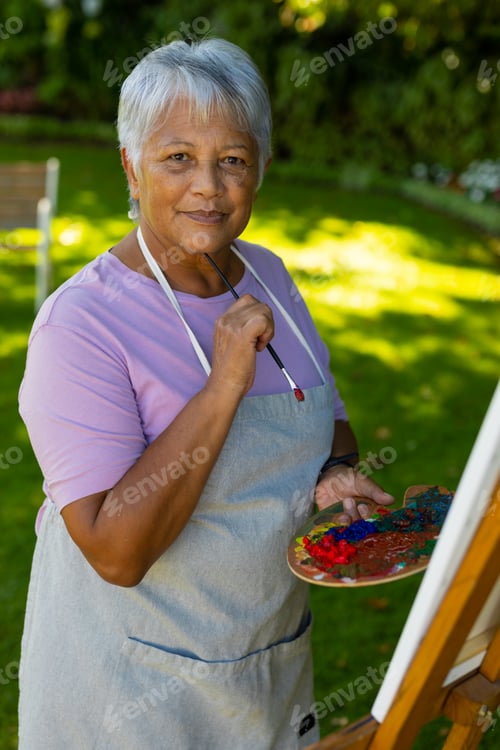 Preview: Portrait of biracial senior woman with short hair wearing apron holding brush and palette in yard