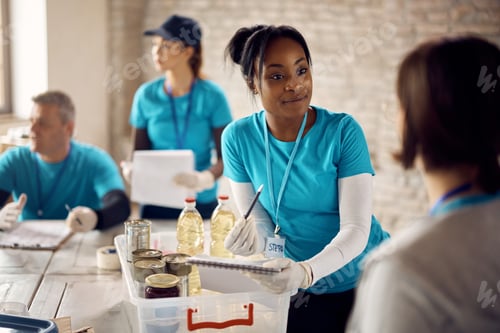 Preview: Young black volunteer going through donation checklist while talking to her associate at food bank.