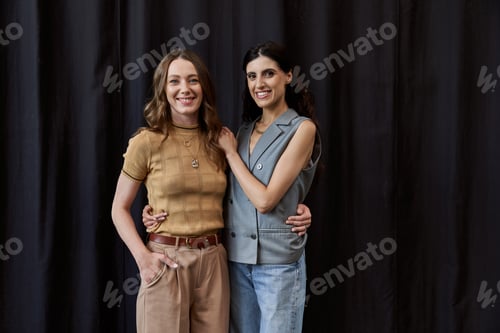 Preview: successful women in stylish casual attire embracing and looking at camera near black drape in office