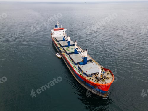 Preview: Large cargo ship with a red bow in open sea, accompanied by a tugboat under an overcast sky