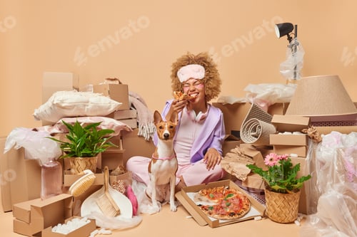 Preview: House property and moving day. Positive young woman with curly hair eats delicious pizza poses near