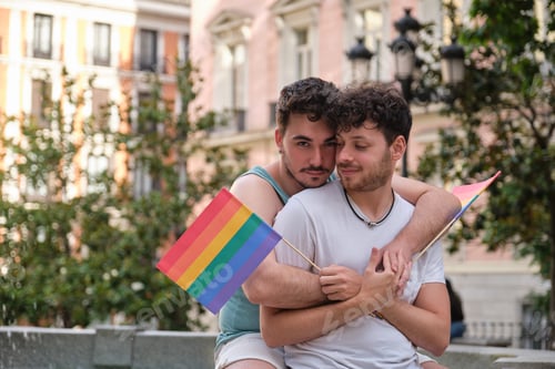 Preview: Gay couple hugging and smiling holding LGBT flags sitting near by a font.