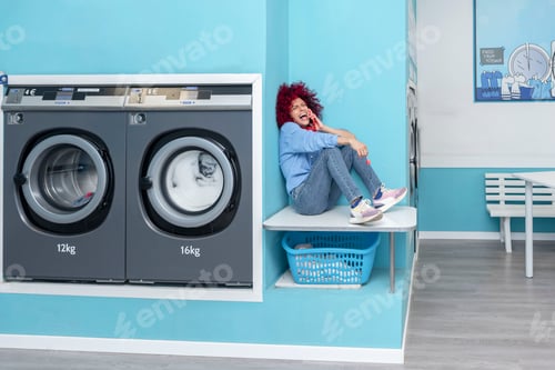 Preview: a laughing young latin woman with afro hair sitting in a laundry room talking on cell phone