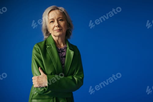 Preview: positive grey haired woman in green leather jacket standing with crossed arms isolated on blue