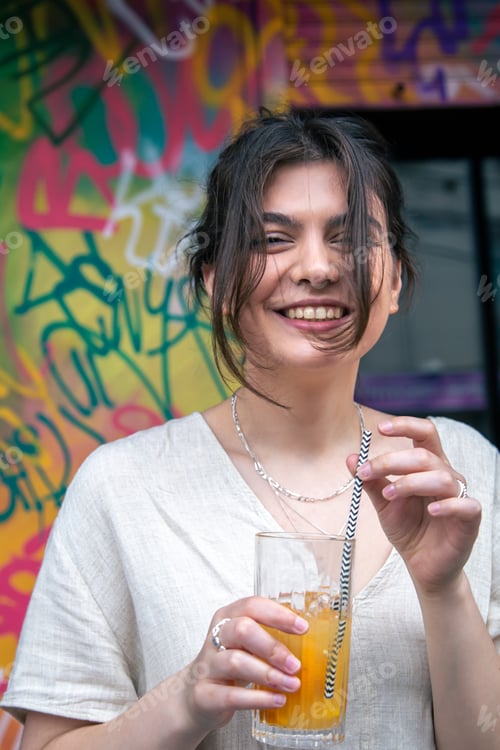Preview: Happy young woman with a glass of lemonade against a bright painted wall.