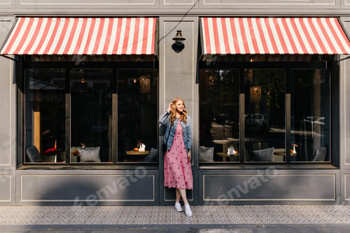 Preview: horizontal full-length photo of a cute curly blonde touching her hair against a street cafe