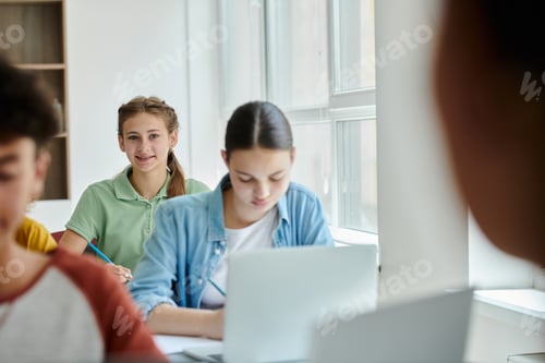 Preview: Teen schoolgirl looking at camera and smiling near blurred classmates during lesson in classroom