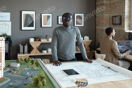 Preview: Young African engineer standing by table with sketch and house layout