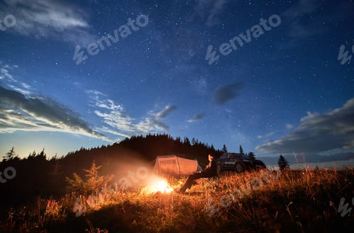 Preview: Young man relaxing next to bonfire on hill.