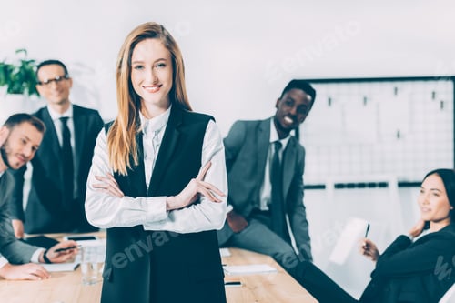 Preview: beautiful young businesswoman looking at camera with blurred multicultural colleagues on background