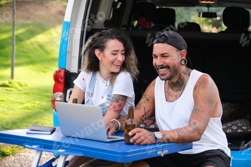 Preview: Young tattooed couple laugh and use the laptop in a fold table near by the van.