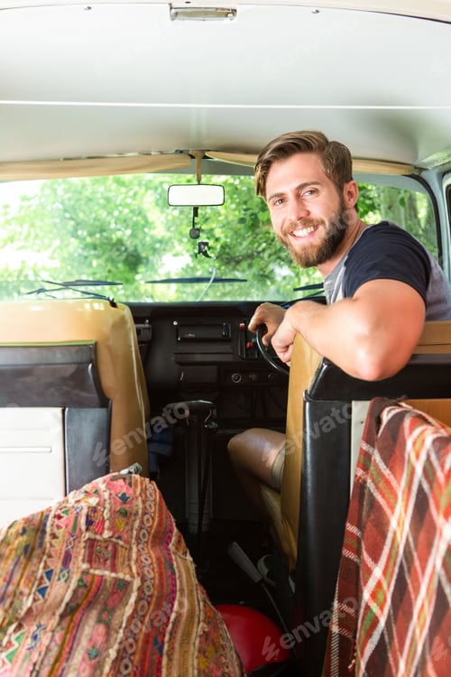 Preview: Man sitting in vintage camper van turning toward camera with action camera in forest, copy space