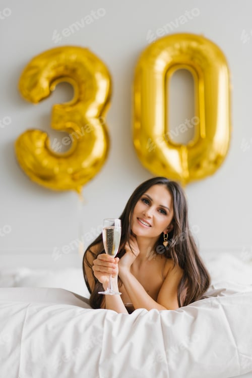 Vista previa: Happy Young Woman Meets her Thirtieth Birthday in bed, Holding a Glass of Champagne in her Hands