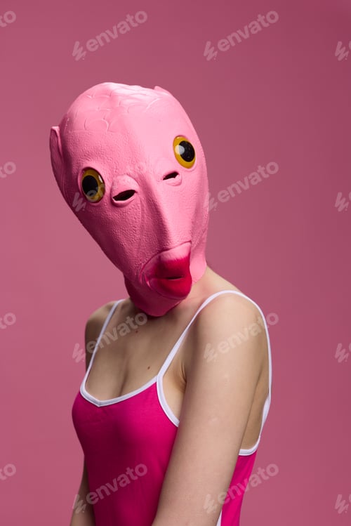 Preview: A woman wearing a pink fish head mask on Halloween stands and looks at the camera against a pink