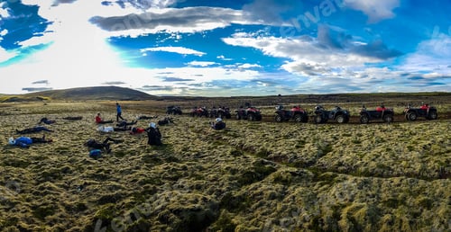 Preview: Panoramic view of people resting near quad bikes in a green field under a cloudy blue sky