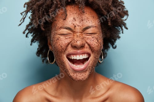Preview: Overjoyed woman laughs loudly, cleans face skin with coffee scrub, has natural mask, crisp hair, pos
