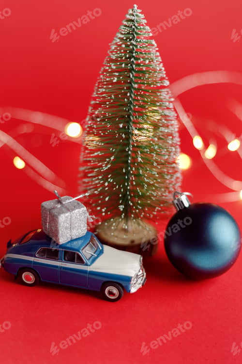 Preview: Toy car with gift box on roof under Christmas tree. New Year and Christmas composition on a red
