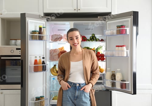 Preview: Happy young woman near open refrigerator in kitchen