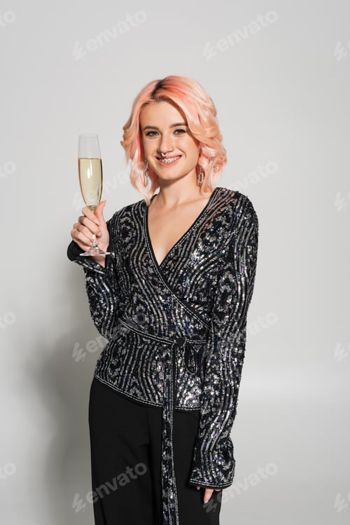 Preview: elegant woman in dental braces smiling at camera and holding champagne glass on grey background