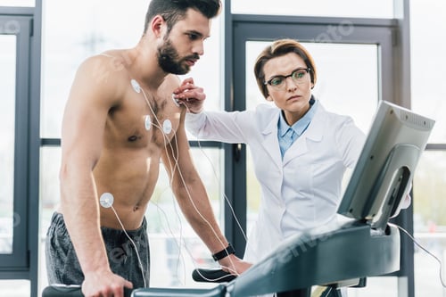 Preview: beautiful doctor putting electrodes on shirtless sportsman during endurance test
