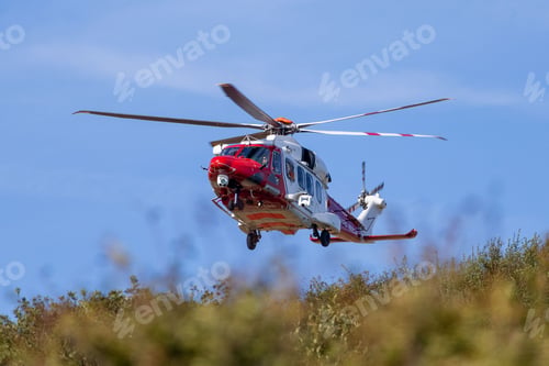 Preview: Rescue helicopter flying over grassy area with clear blue sky.