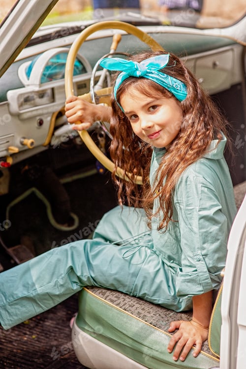 Preview: Cute little girl in a blue overalls and a scarf on her head, sits behind the wheel in a retro car