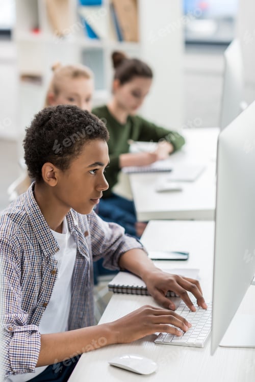 Preview: side view of african american teen boy typing on keyboard in front of computer screen in class