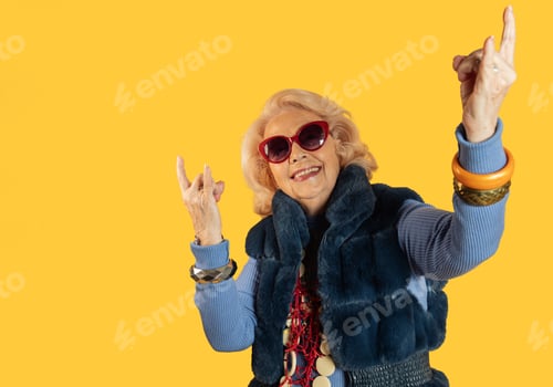 Preview: Stylish senior woman making rock and roll hand gesture on yellow background
