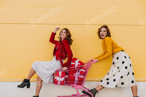 Preview: Graceful girl using shopping cart. Two happy friends having fun while posing on the street