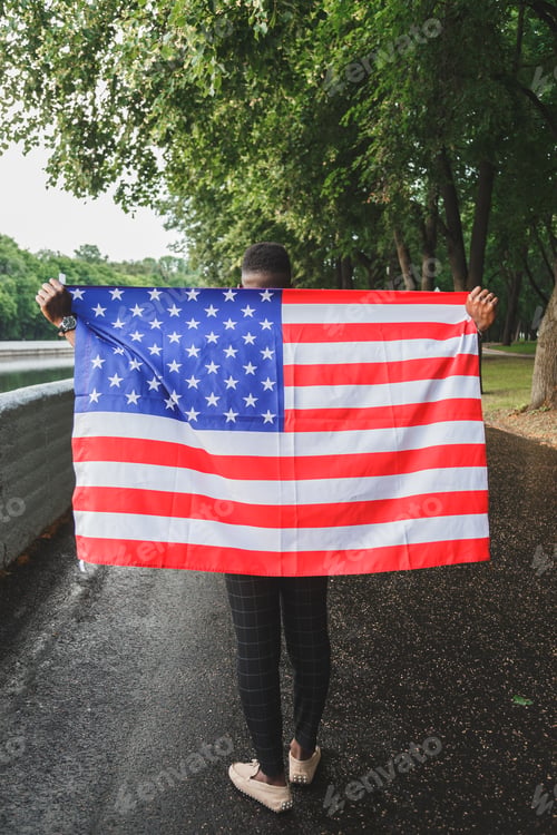 Preview: Black african-american man holding USA flag from his back walking outdoors