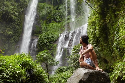 Preview: Tourism, travel and adventure. Stylish young hipster sitting on stone with bare feet and turning his