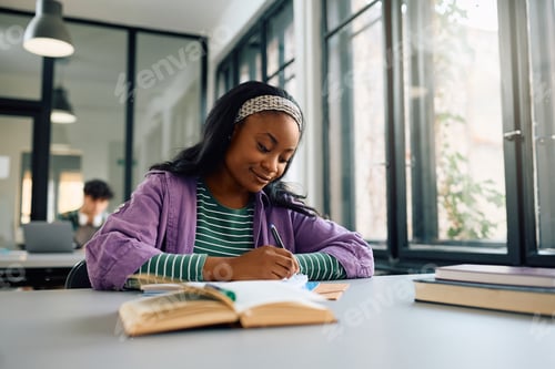 Preview: Black female student writing notes while learning in a library.