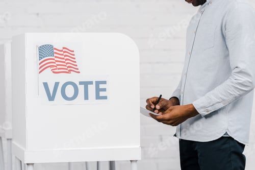 Preview: cropped view of african american citizen holding pen and ballot while voting near stand with vote