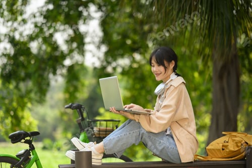 Preview: Smiling Asian female student using laptop, sitting on wooden bench in the park