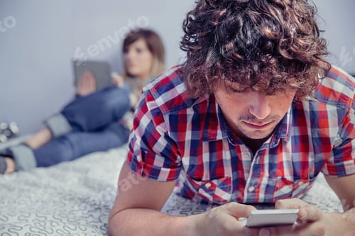 Preview: Man using smartphone over bed and woman reading in tablet