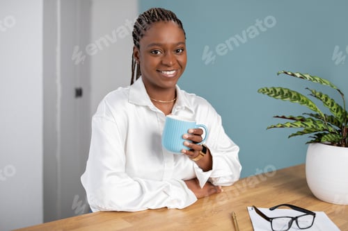 Preview: Positive young black woman sitting at workdesk, drinking coffee
