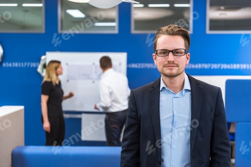 Preview: Businessman standing in conference room, while colleagues working with flipchart in background
