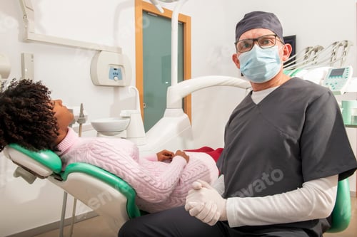 Preview: Dentist wearing surgical mask posing next to patient lying on dentist chair