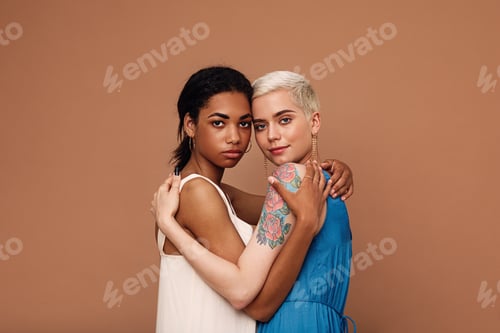 Preview: Two beautiful women embracing in the studio. Females in dresses looking at camera.