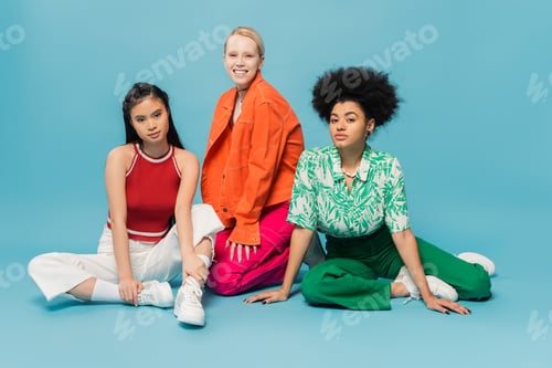 Preview: full length of young multicultural women in bright and stylish attire sitting on blue background