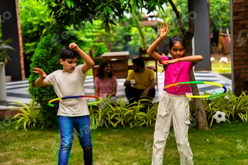 Preview: Smiling Indian parents watching kids play hula hoop in backyard garden on a sunny day